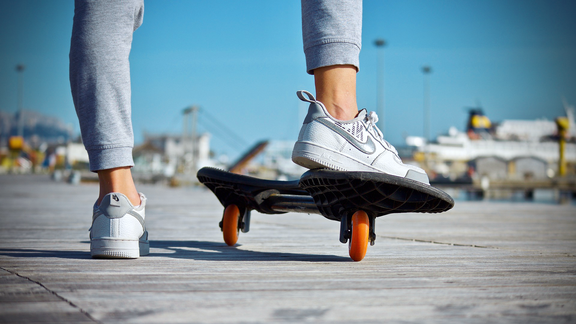 Zdjęcie przedstawia nogi osoby od kolan w dół. Osoba ubrana jest w spodnie dresowe i sportowe buty. Prawa noga oparta jest na deskorolce na dwóch kółkach.