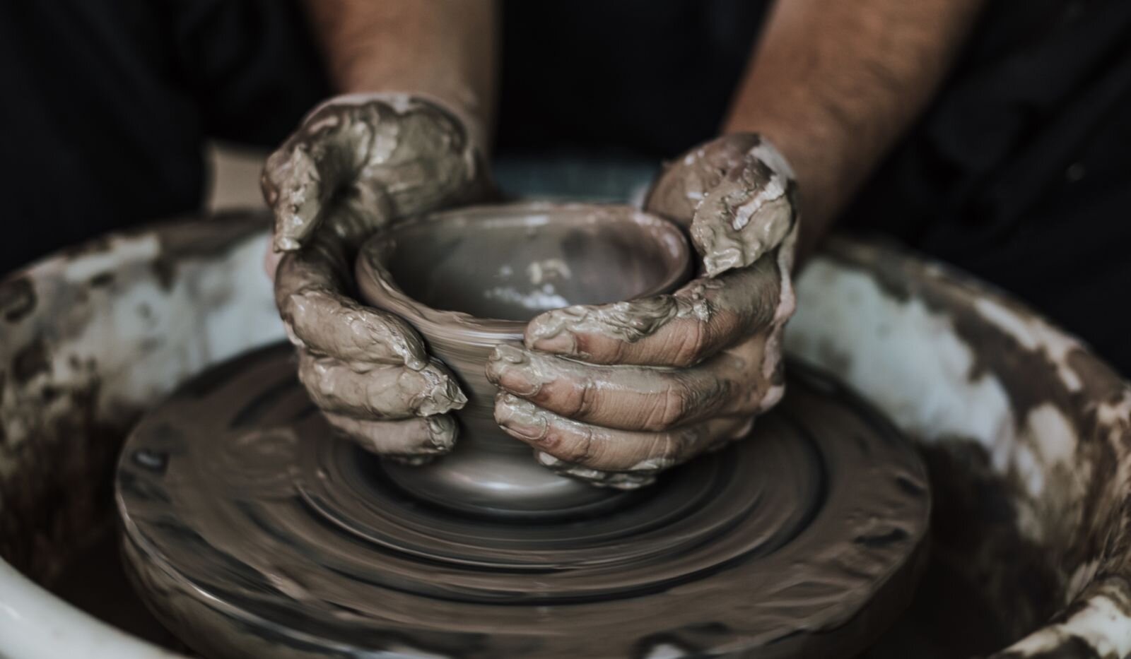 Zdjęcie przedstawia zbliżenie na ręce garncarza toczącego naczynie z gliny na kole garncarskim. Glina jest rzadka i przecieka przez palce, które umiejętnie formują mały, prosty wazon.