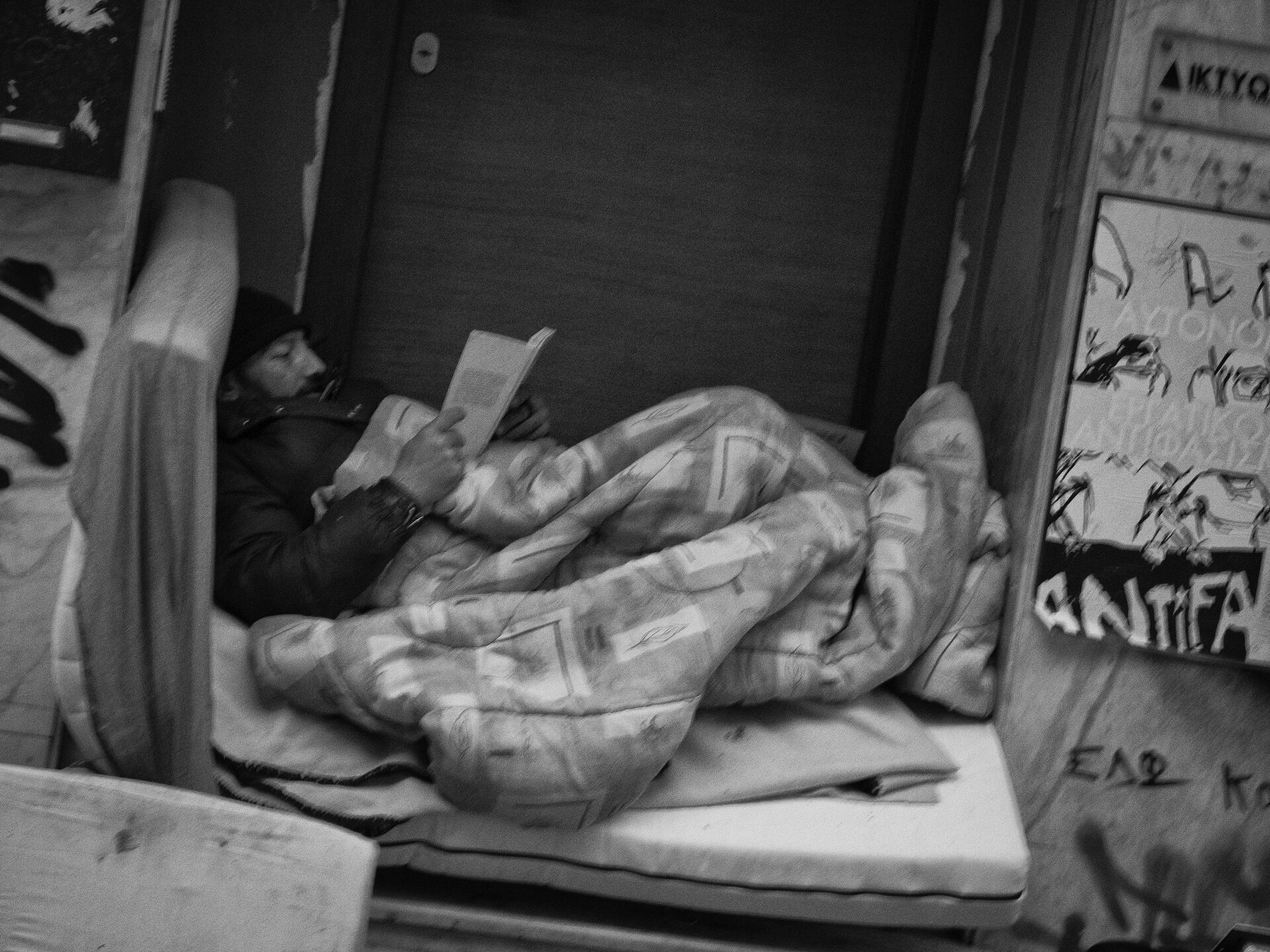 Ilustracja przedstawia osobę bezdomną. Postać ubrana w kurtkę oraz czapkę, leży na materacu, przykryta kołdrą i czyta książkę.