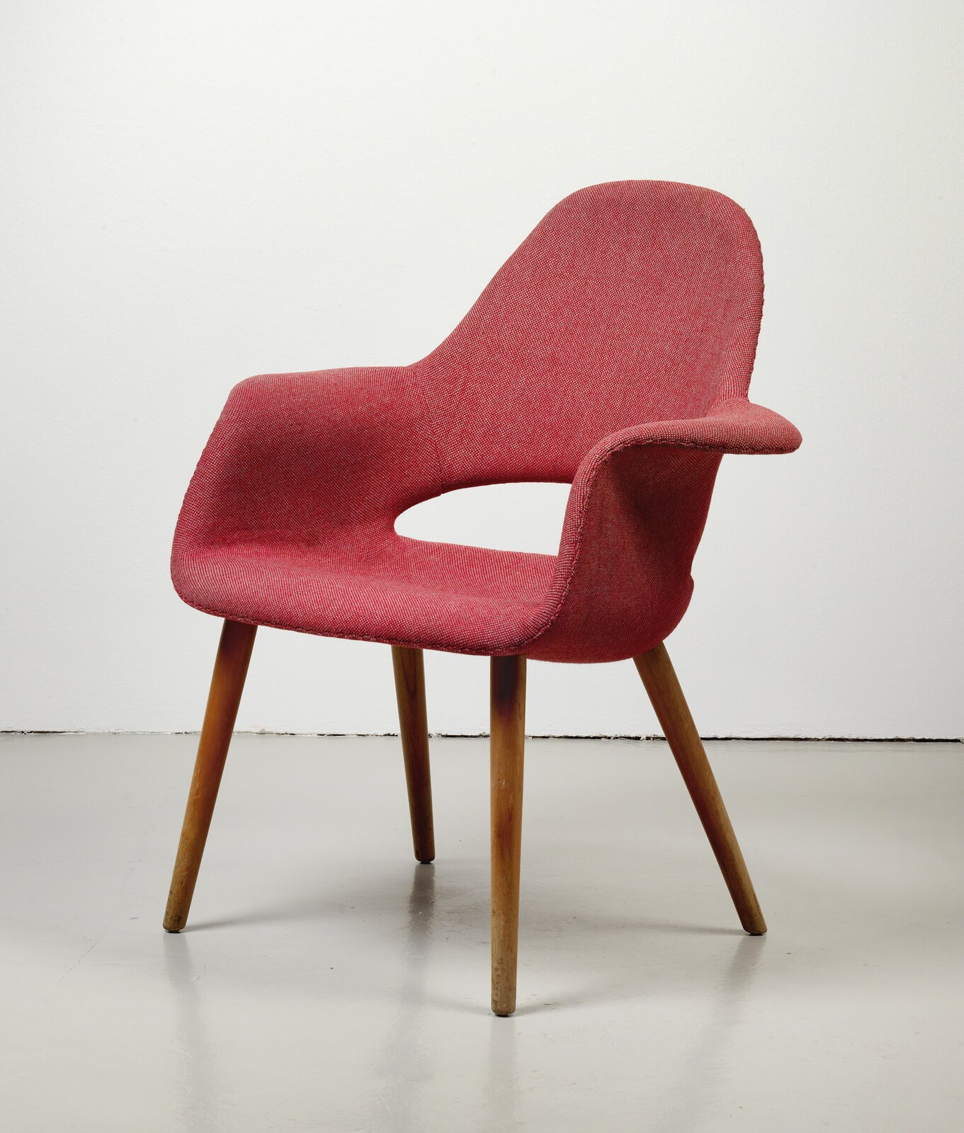 Fotografia przedstawia czerwone krzesło stojące na czterech drewnianych nogach. Krzesło ma czerwone obicie, i otwór pomiędzy siedziskiem a oparciem. Z prawej strony krzesła - podłokietnik.