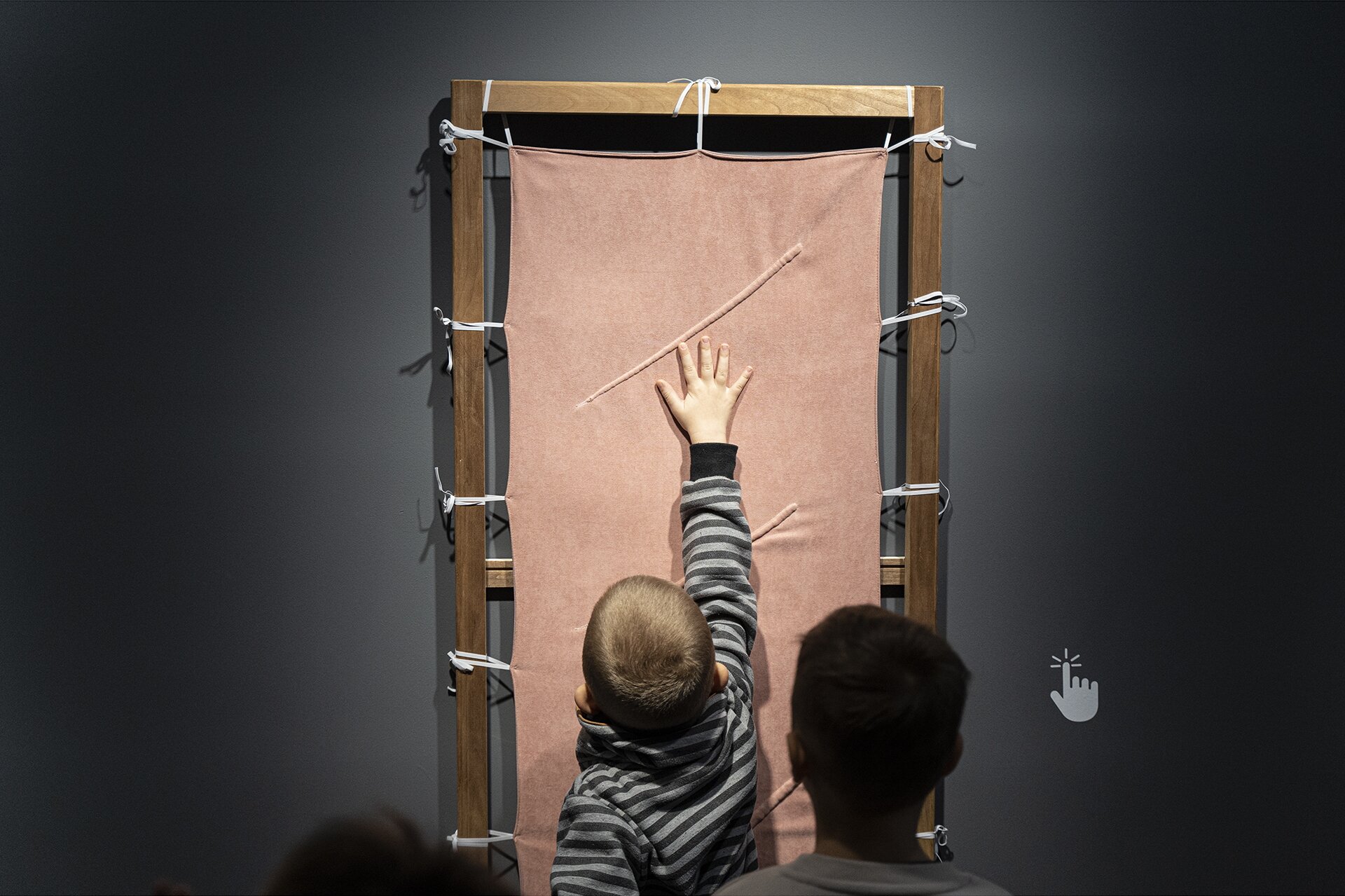 Fotografia ukazuje dwóch chłopców zwróconych tyłem do widza. Na szarej ścianie ukazana jest kompozycja składająca się z tkaniny przywiązanej białym sznurkiem do drewnianej ramy. Jeden z chłopców, prawą ręką dotyka tkaniny. 