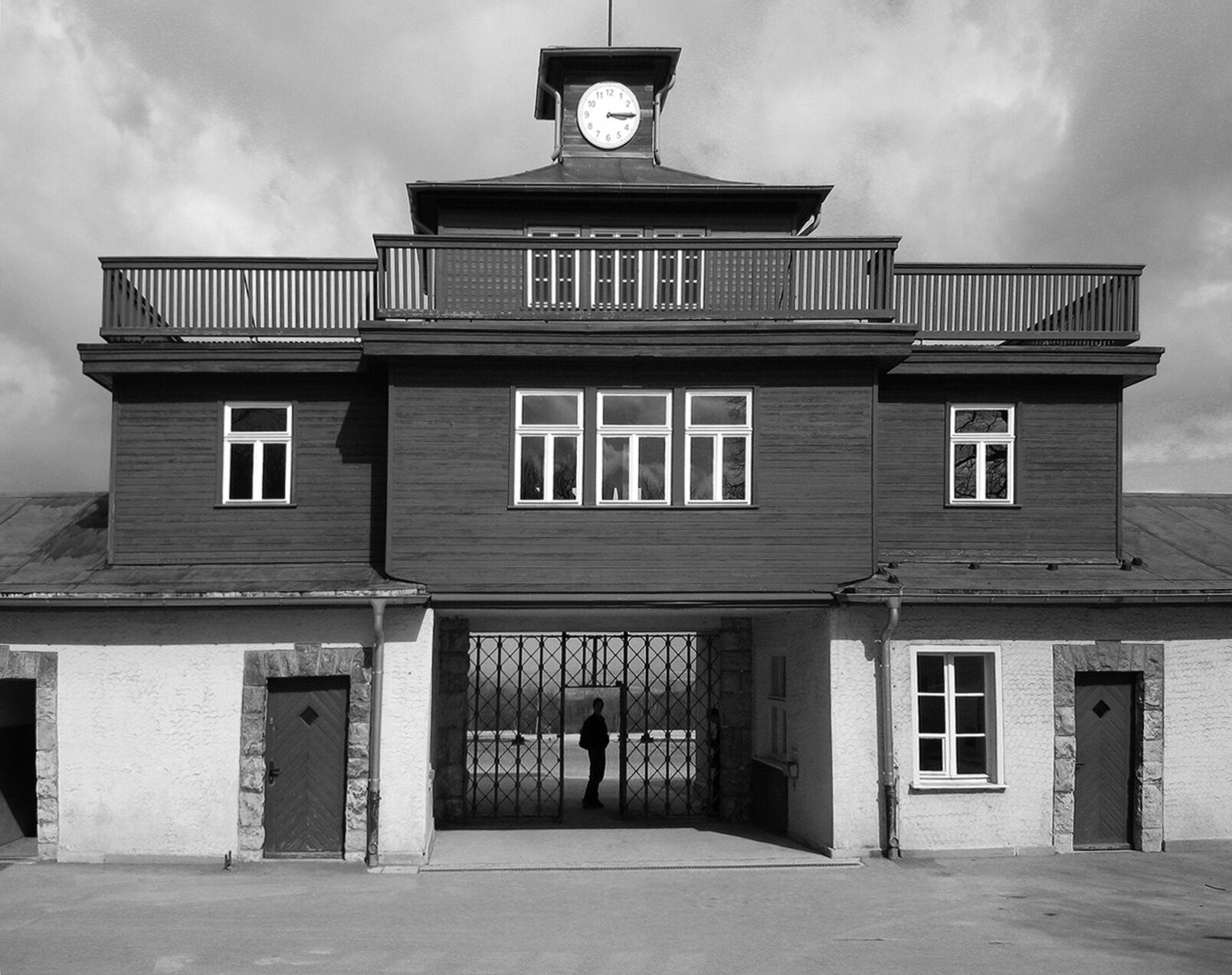 Fotografia czarno-biała przedstawiająca bramę obozu Buchenwald. To budynek do połowy murowany z  dwoma wejściami, na górze drewniany. Zakończony wieżą w zegarem.