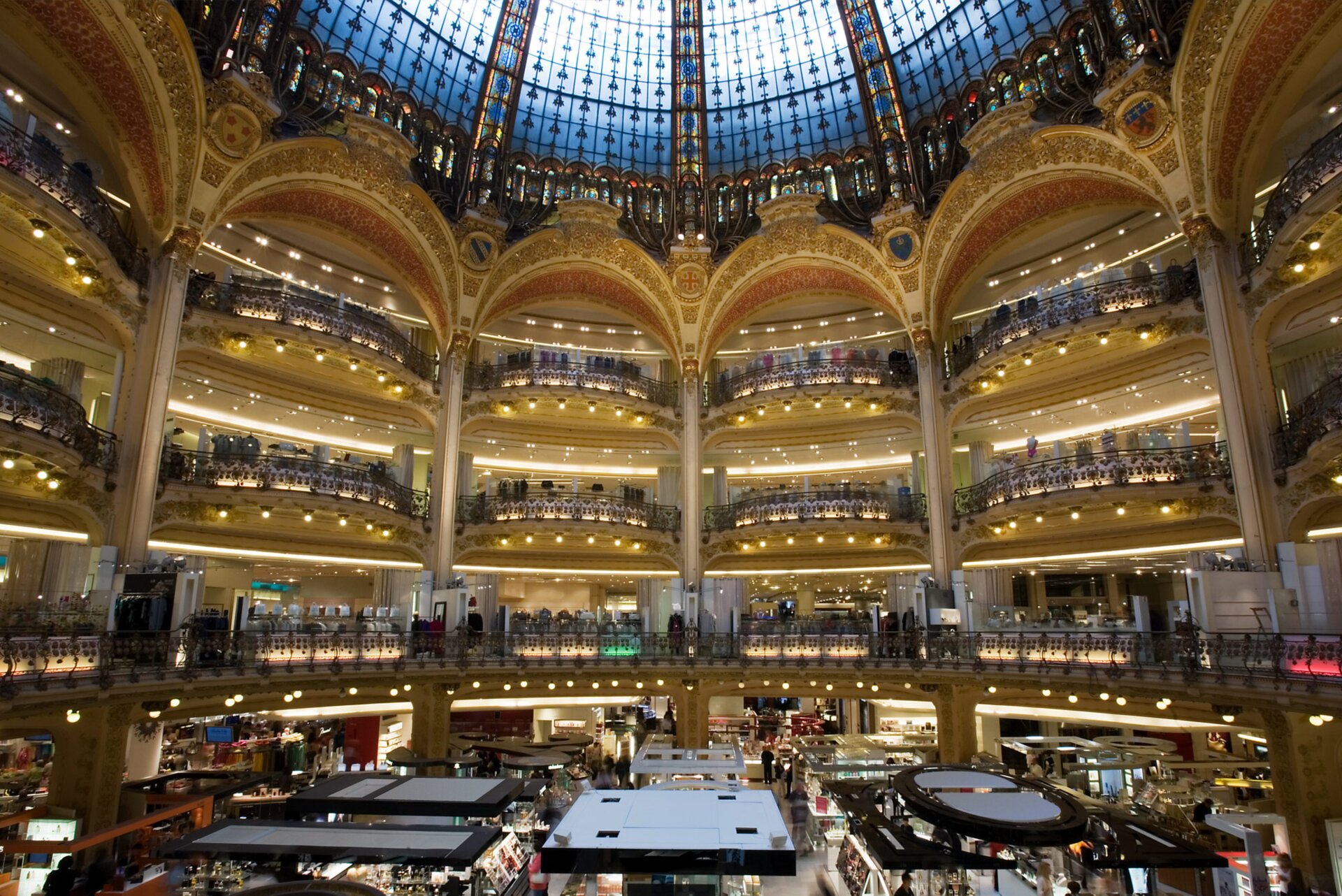 Zdjęcie przedstawia wnętrze wielopiętrowego, zabytkowego budynku galerii handlowej. Widoczna jest wysoka, okrągła hala, zakończona ażurową kopułą wykonaną z witrażowych szyb, poniżej której widoczne są trzy piętra z pasażami, które od strony wewnętrznej są zakończone balkonami zwieńczonymi ozdobnymi łukami. Piętra są wizualnie połączone kolumnami podpierającymi łuki nad ostatnim piętrem. Na parterze widoczne są stoiska z produktami. Całość oświetlona jest dużą ilością ciepłego światła. Na każdym pietrze i na parterze widoczni są ludzie.