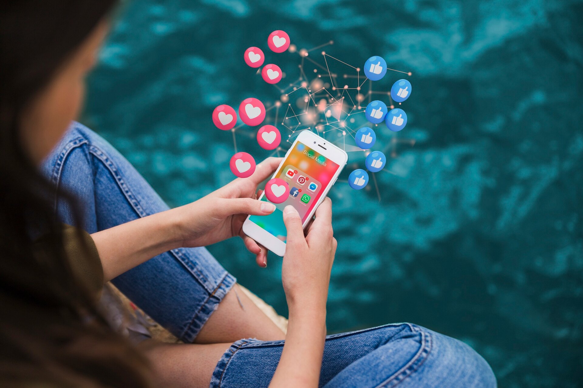 Zdjęcie przedstawia dziewczynę siedzącą na brzegu basenu i oglądającą coś na smartfonie. Ze smartfona sypią się lajki i serduszka.