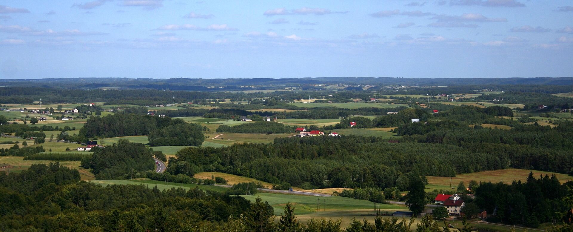 Zdjęcie przedstawia Pojezierze Pomorskie, na ilustracji ukazane zostały tereny trawiaste mieszające się z terenami zalesionymi.  W niektórych miejscach znajdują się budynki i drogi. 