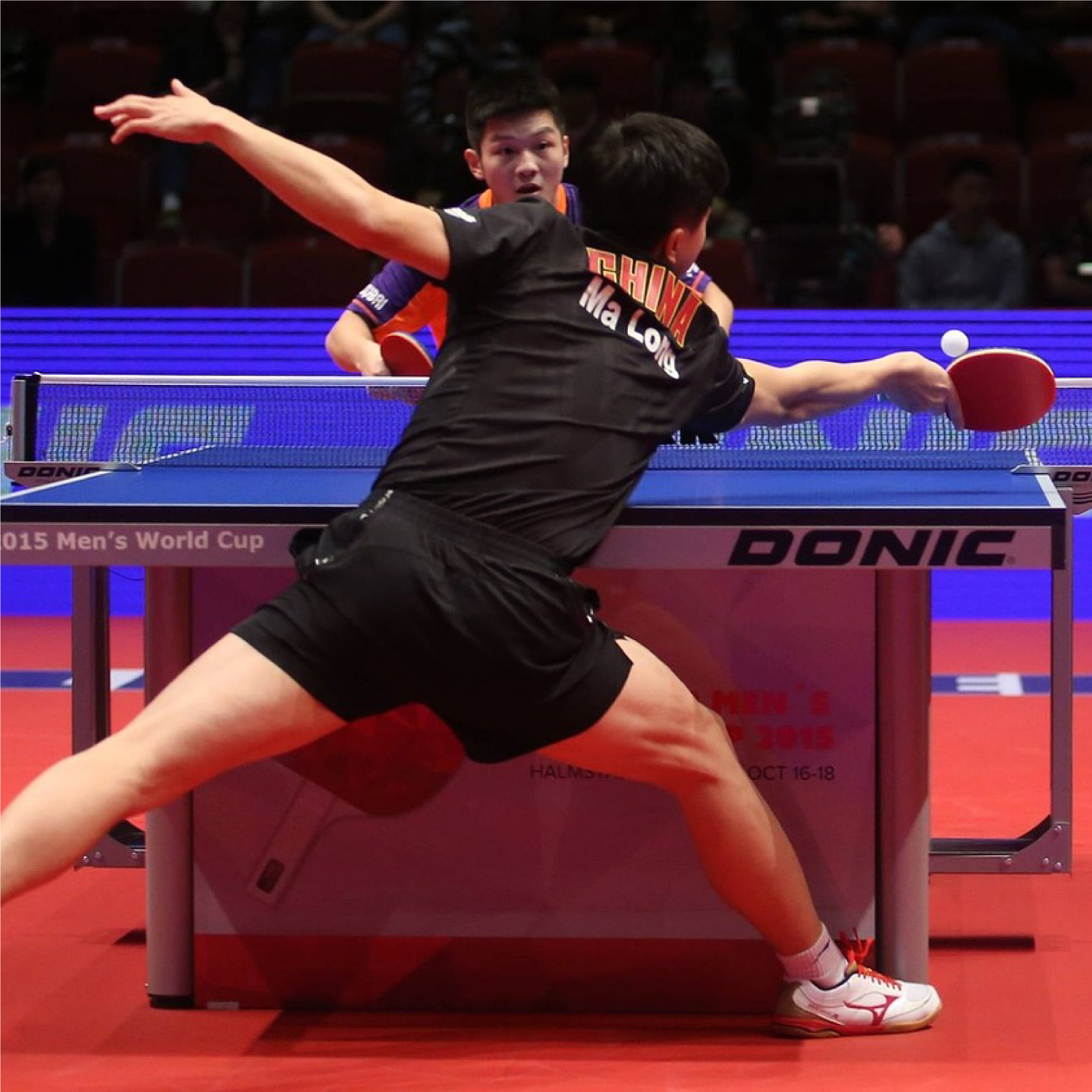 Zdjęcie przedstawia dwóch mężczyzn grających w ping‑ponga na hali sportowej z pełną widownią.