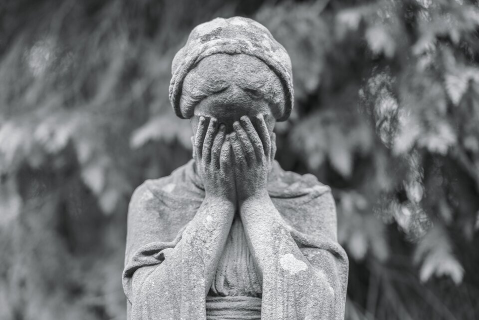 Zdjęcie przedstawia fragment pomnika nagrobnego. Widać tu postać kobiety z opuszczoną głową, zasłaniającej dłońmi twarz.