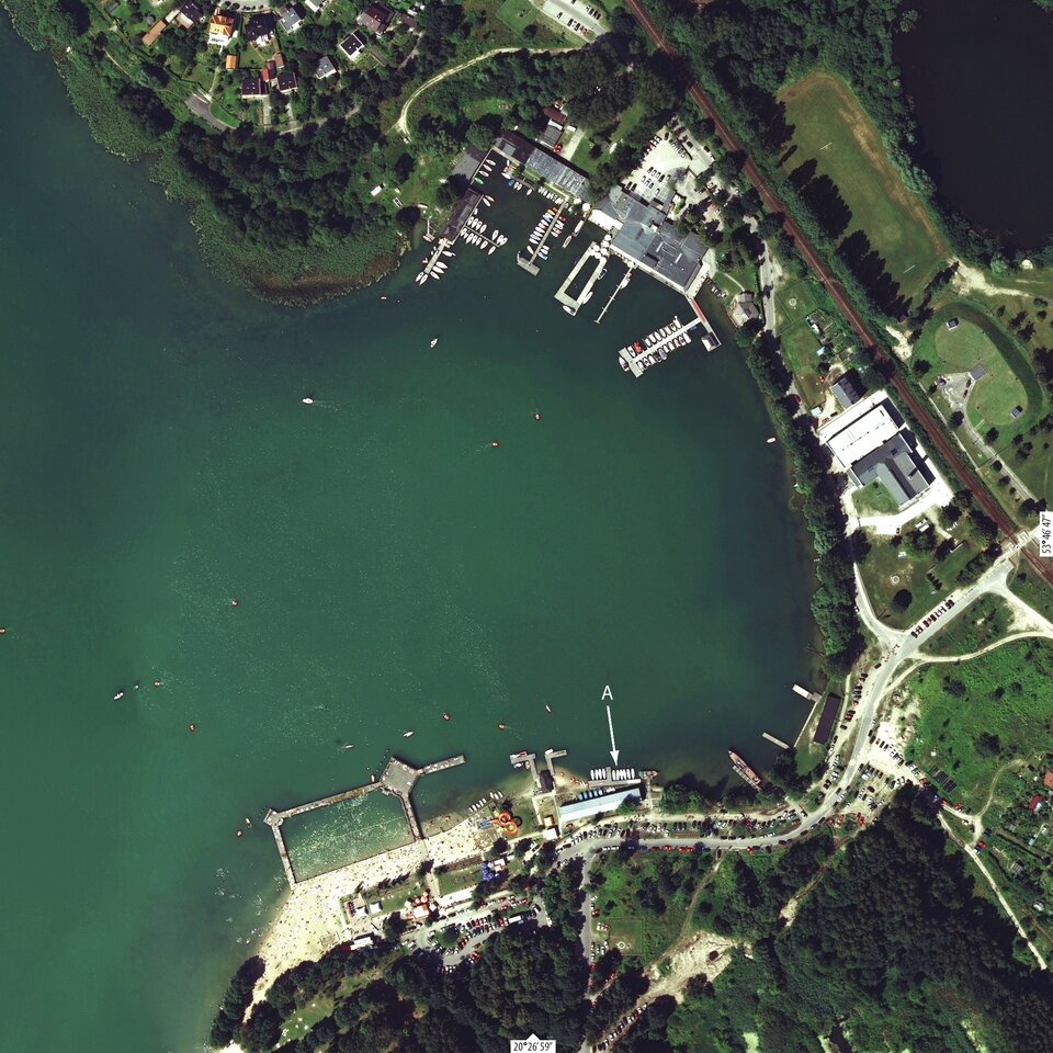 Zdjęcie satelitarne przedstawia fragment jeziora Ukiel w Olsztynie. Jezioro ma zaokrąglone brzegi. Na brzegu znajduje się molo, plaże oraz budynki.