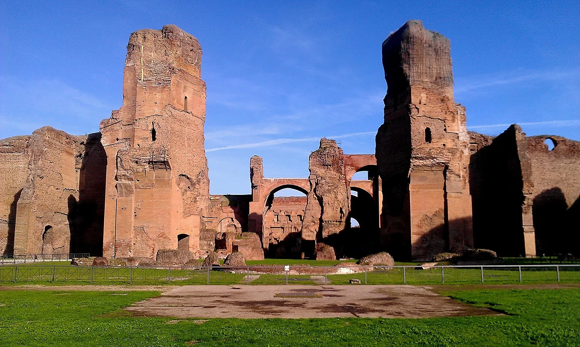 Widok na Termy Karakalli - kompleks monumentalnych budowli, złożonych z kolumn, łuków, brył. Przed kompleksem zielona trawiasta polana, nad nim - niebieskie niebo.