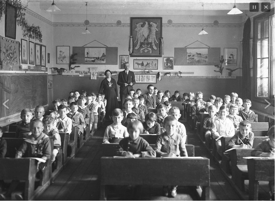 Fotografia przedstawia klasę pełną uczniów siedzących w ławkach patrzących w obiektyw. Z tyłu klasy widać dwoje nauczycieli, kobietę i mężczyznę. Na ścianach wiszą obrazki, po środku klasy najwyżej wisi godło Polski, orzeł. Okna w sali są otwarte, klasa jest bardzo liczna.