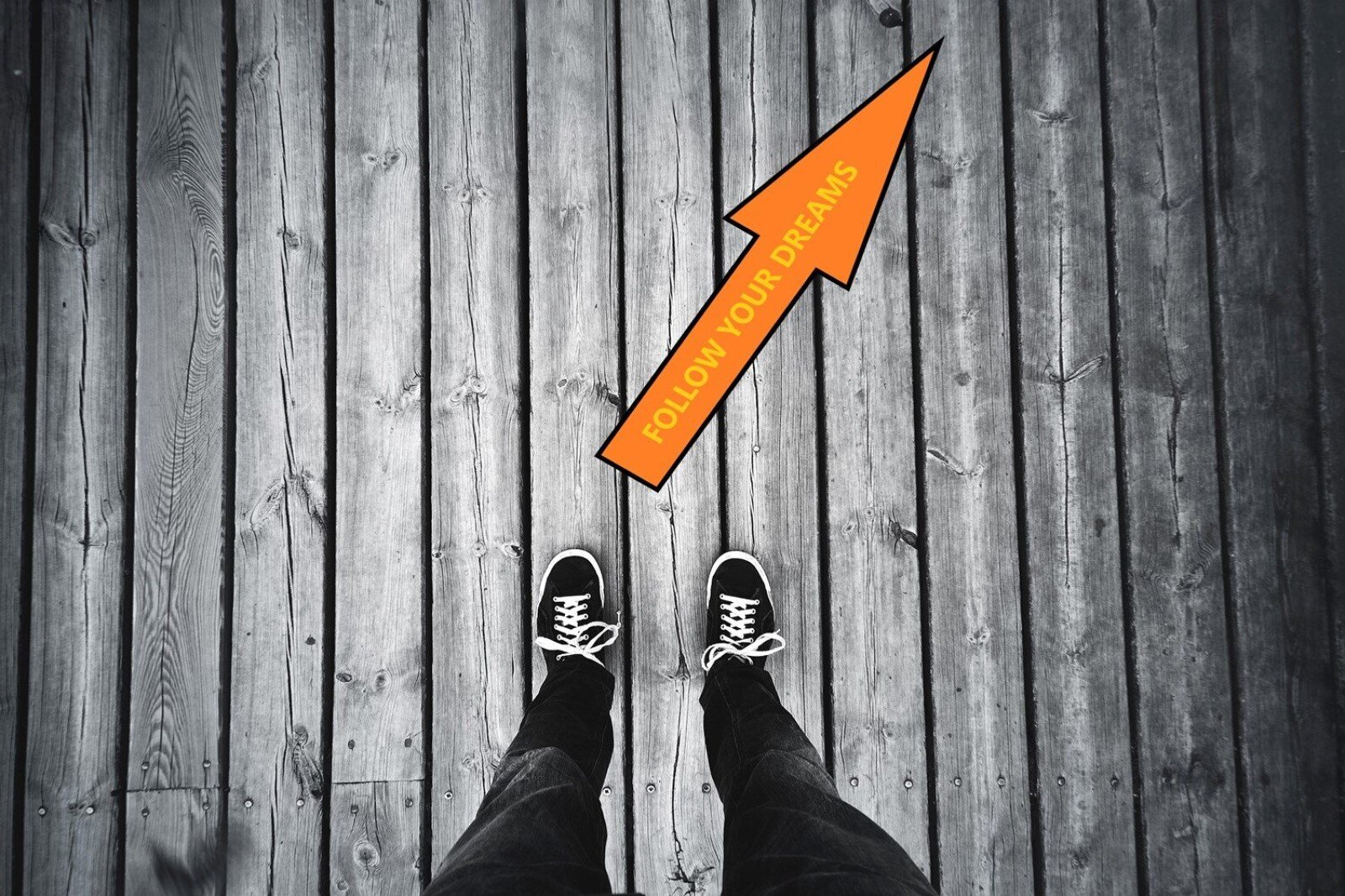 Zdjęcie przedstawia ciemną, drewnianą podłogę z lotu ptaka oraz męskie nogi w dżinsach i trampkach. Przed nimi pomarańczowa strzałka z żółtym napisem  Follow your dreams