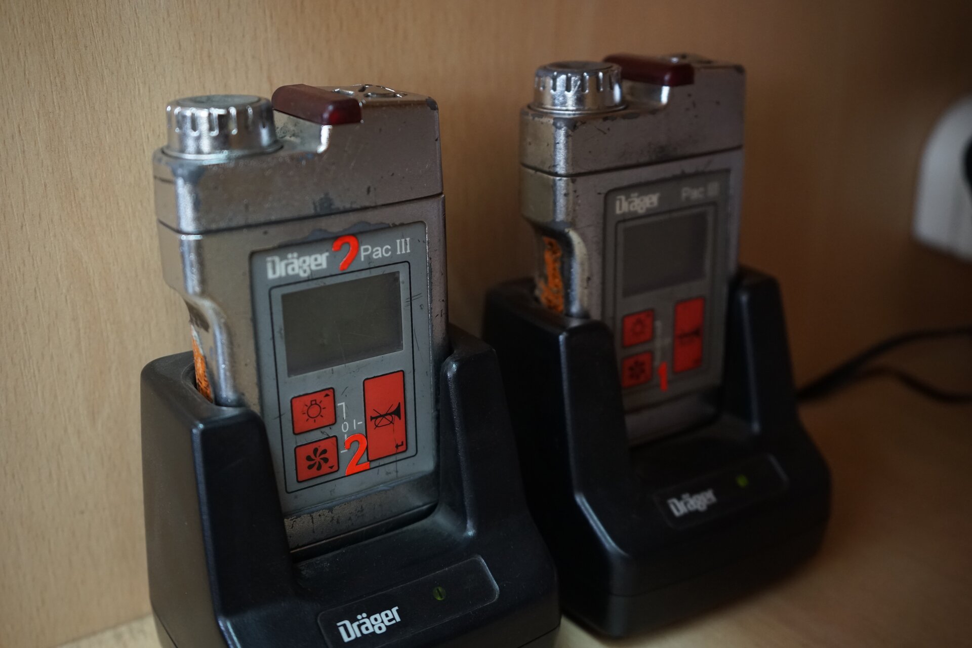 Zdjęcie przedstawia dwa detektory gazu przeznaczone do wykrywania tlenku węgla. Detektory są urządzeniami o kształcie prostopadłościanu. Na górnej podstawie detektora widoczne jest srebrne pokrętło. Pokrętło ma cylindryczny kształt. Jego górna część jest wyfrezowana. Z przodu detektora znajduje się wyświetlacz. Pod nim zlokalizowane są trzy przyciski. Po lewej - przycisk z symbolem światła oraz wentylatora. Po prawej przycisk z przekreśloną trąbką. Detektory znajdują się we wsuwanych ładowarkach.