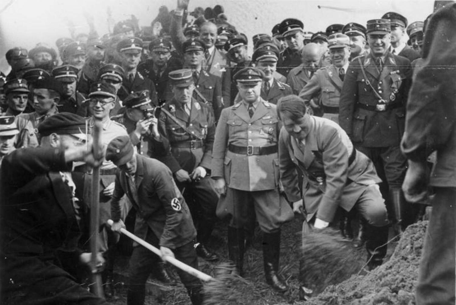 Zdjęcie przedstawia plac budowy. Znajduje się na nim duża grupa żołnierzy niemieckich. Wśród nich jest Adolf Hitler z łopatą pełną ziemi.