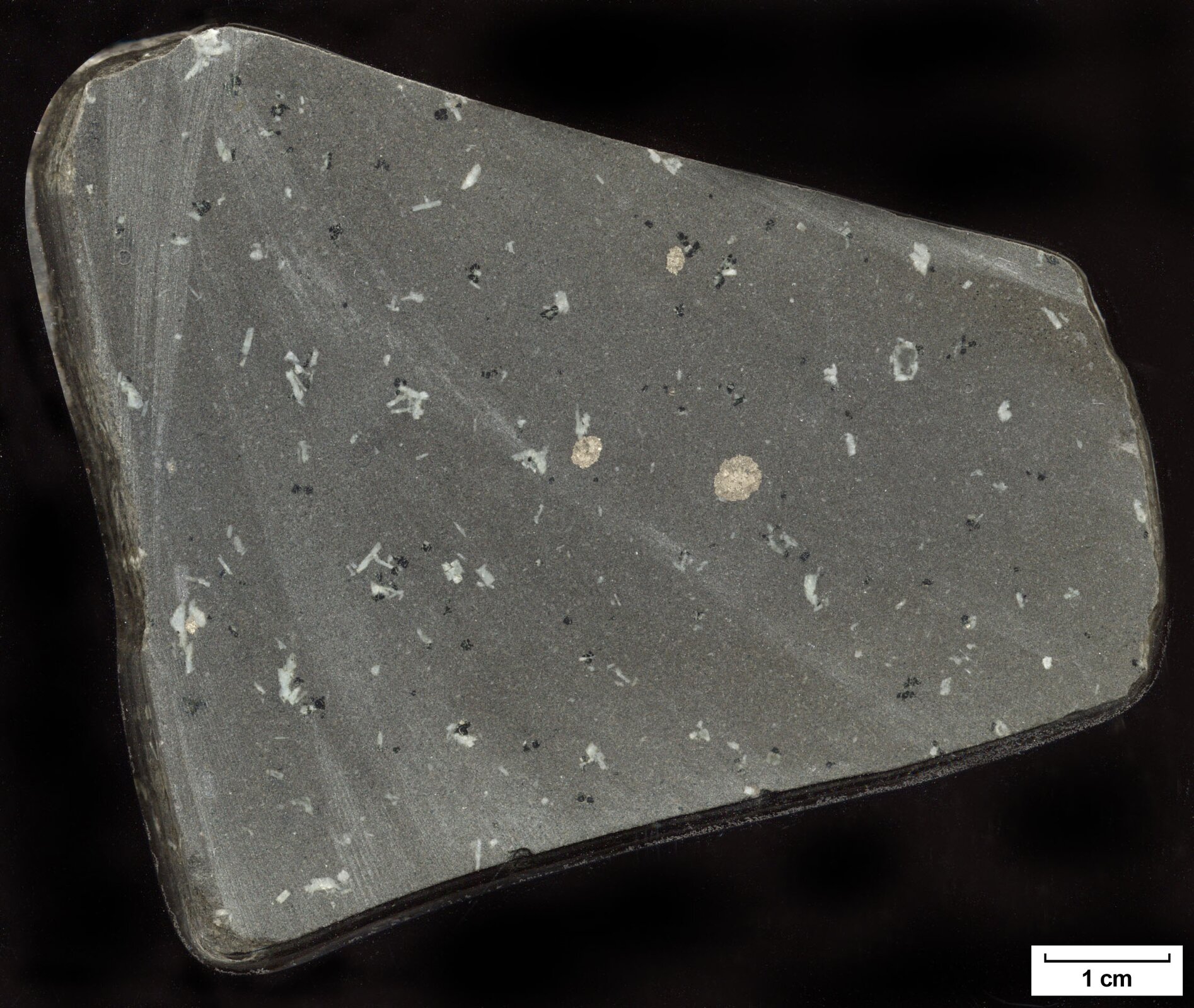 Na zdjęciu przedstawiony jest bazalt – skała lita o strukturze bardzo drobnoziarnistej i barwie ciemnoszarej. W drobnoziarnistej masie skalnej małe kryształy jaśniejszego koloru. Fragment skały przypomina trapez, jest płaski i ma wielkość osiem na sześć centymetrów.
