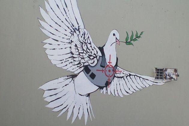 Ilustracja przedstawia białego gołębia, który w dziobie trzyma gałązkę oliwną. Gołąb ubrany jest w kamizelkę kuloodporną. Znajduje się na niej czerwony symbol celowania z broni. 