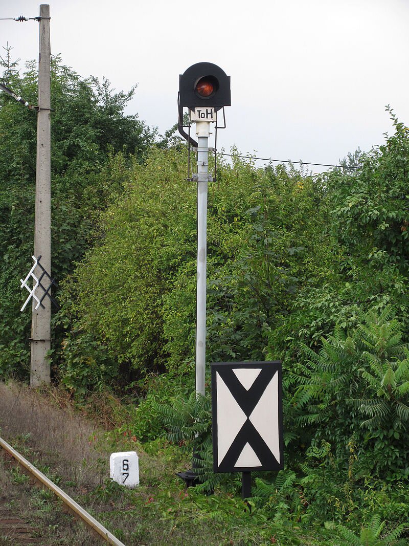 Zdjęcie przedstawia jednokomorową tarczę ostrzegawczą semafora wjazdowego przed tarczą wskaźnik W1. Tarcza umiejscowiona jest przy torach, w tle widać dużo zieleni.  
