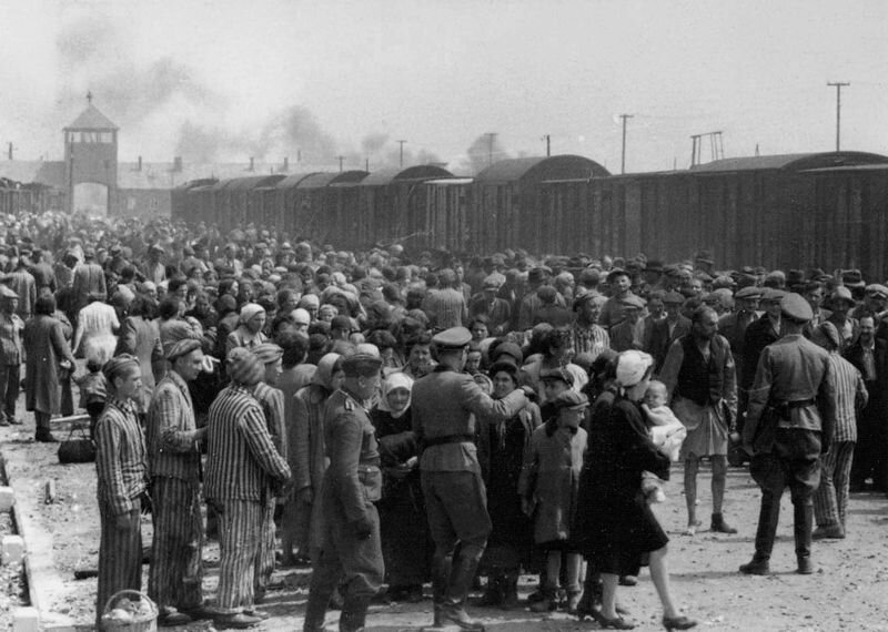 Zdjęcie przedstawia rampę kolejową, na której są tłumnie zgromadzeni ludzie. Wśród nich chodzą niemieccy żołnierze. Części ludzi ubrana jest w cywilne ubrania, część ma na sobie obozowe pasiaki. Na drugim planie stoją towarowe wagony i znajduje się brama wjazdowa. Na niebie widoczne unoszące się dymy.