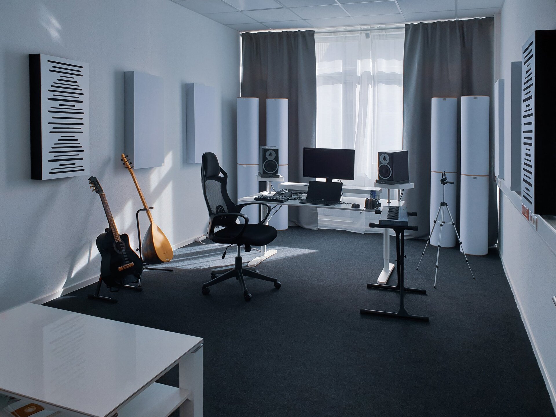 Pomieszczenie na zdjęciu ma jasne ściany i sufit oraz grafitową podłogę. W lewym dolnym rogu znajduje się biały niski stolik z półką pod blatem. Na środku jest czarne krzesło na kółkach, keyboard na czarnym stojaku oraz białe biurko. Na biurku leży czarny laptop, za nim na podwyższeniu stoi czarny monitor. Po bokach monitora, również na podwyższeniu, stoją kolumny głośnikowe w kształcie prostopadłościanu. Mają po dwa okrągłe głośniki. Dolny głośnik jest czarny z jasną ramką, a górny jasny z małym, czarnym trójkątem w środku. Po lewej stronie laptopa leżą dwie małe konsole muzyczne i kable. Po prawej stronie jest myszka na kablu, czarne słuchawki oraz niebieska puszka. Za krzesłem stoją oparte o czarne stojaki dwa instrumenty. Pierwszy to czarno‑brązowa gitara, a drugi to mandolina w kolorze jasnego drewna.  Po prawej stronie biurka stoi srebrno‑czarny trójnogi statyw. W tle znajduje się duże okno z białą firanką i szarymi zasłonami po bokach. Przed zasłonami stoją po dwie duże, białe kolumny. Na ścianach są powieszone po trzy panele w kształcie prostopadłościanu. Jeden biały z czarnymi bokami i czarnymi, poziomymi liniami o różnej długości oraz dwa całe białe.  