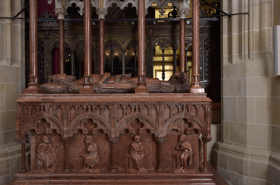 Zdjęcie przedstawia nagrobek Kazimierza Wielkiego. Jest on cały drewniany. Na wieku nagrobka znajduje się figura władcy w szatach leżącego na poduszce. Nagrobek jest ozdobiony łukami oraz figurami osób. W górę odchodzą kolumny.