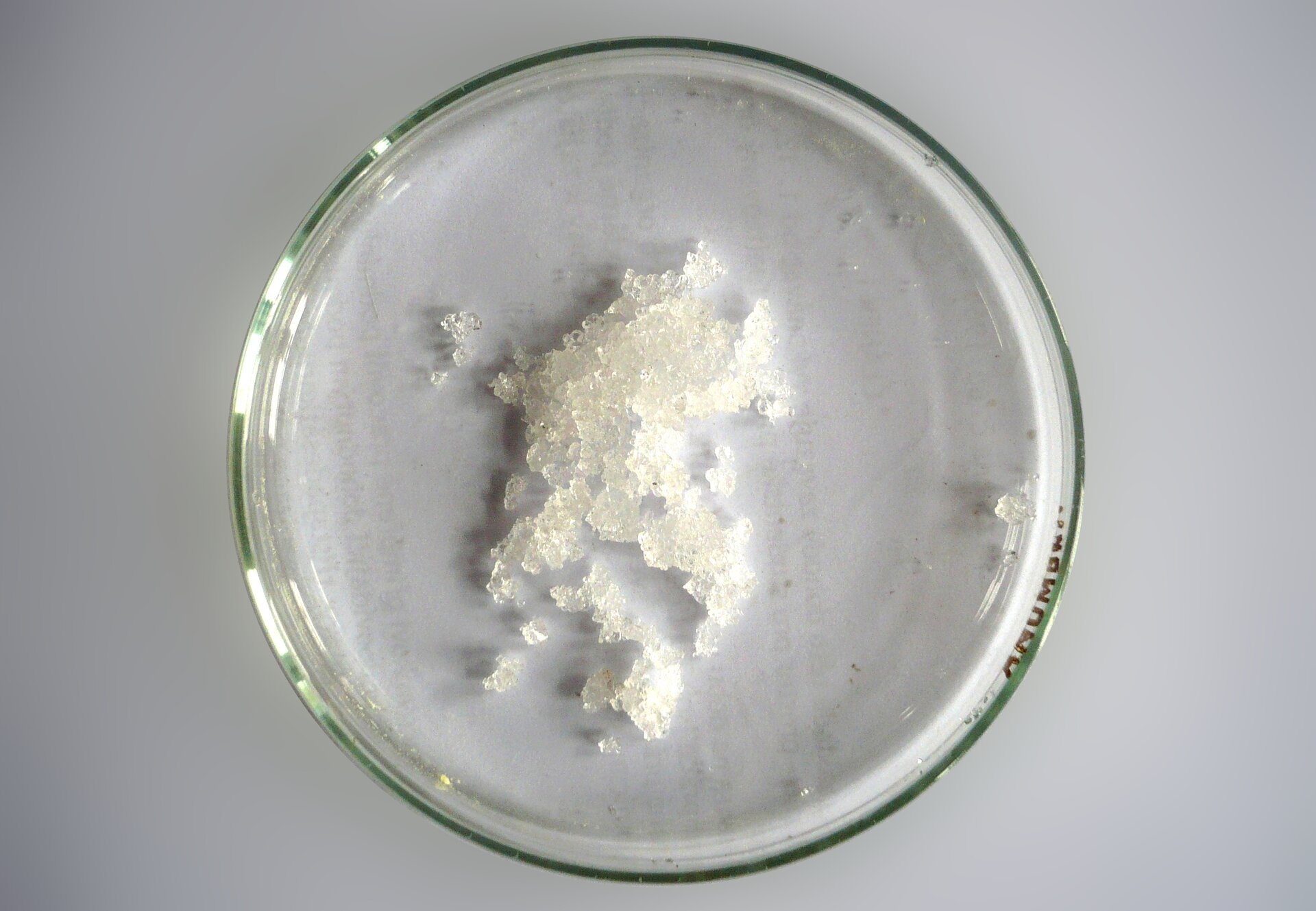 Zdjęcie przedstawiające szalkę Petriego oglądaną od góry. W naczyniu znajduje się niewielka porcja białych, pozlepianych ze sobą kryształków. Jest to chlorek magnezu.