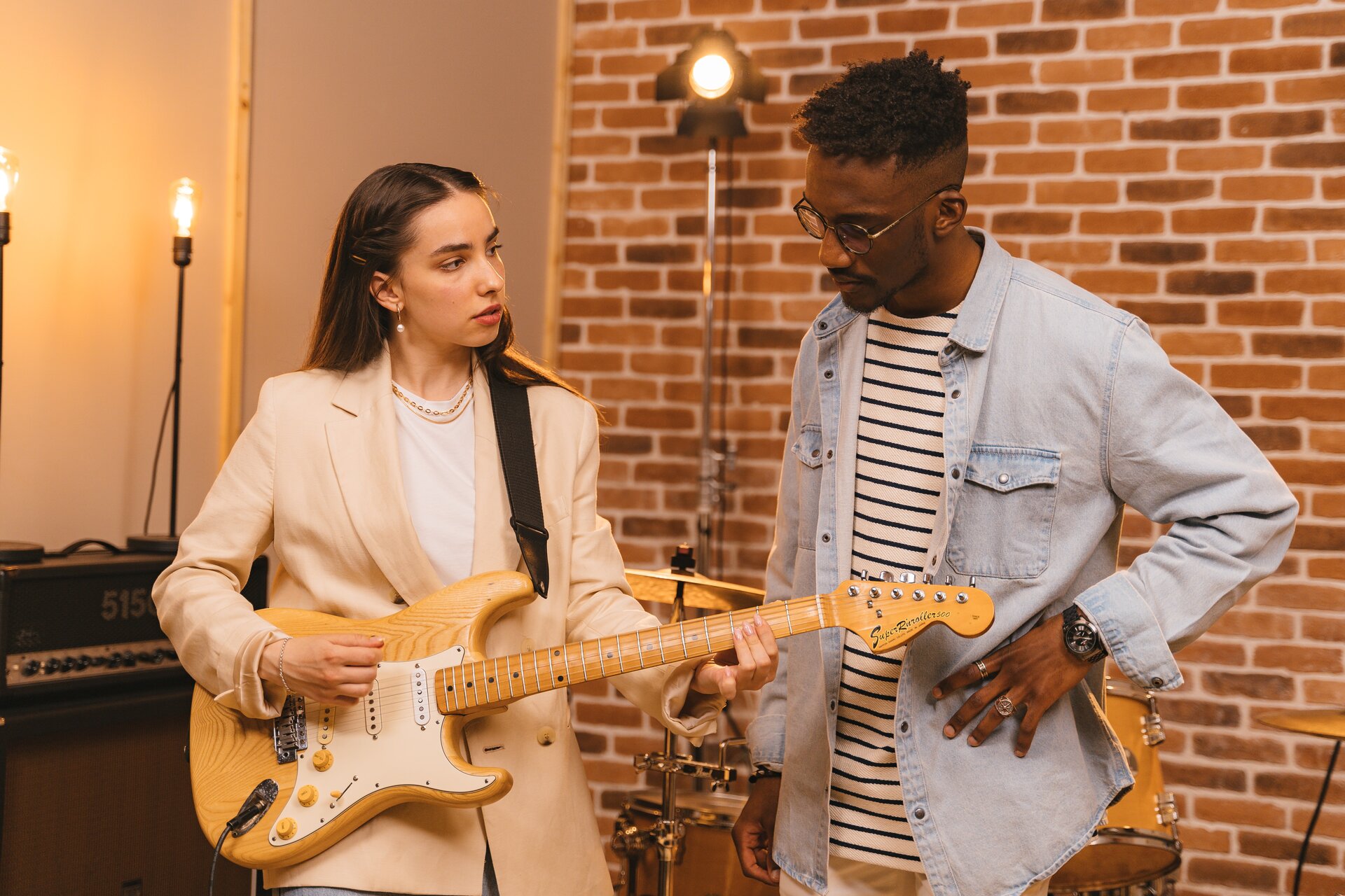 Zdjęcie przedstawia młodą kobietę trzymającą gitarę. Obok niej stoi młody mężczyzna i przygląda się jak kobieta układa dłoń na strunach.