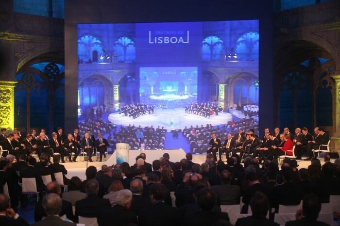 Zdjęcie przedstawia dużą salę,  w której zasiadają licznie mężczyźni i kobiety. W tle zdjęcie tej samej sali z napisem w języku portugalskim Lizbona.