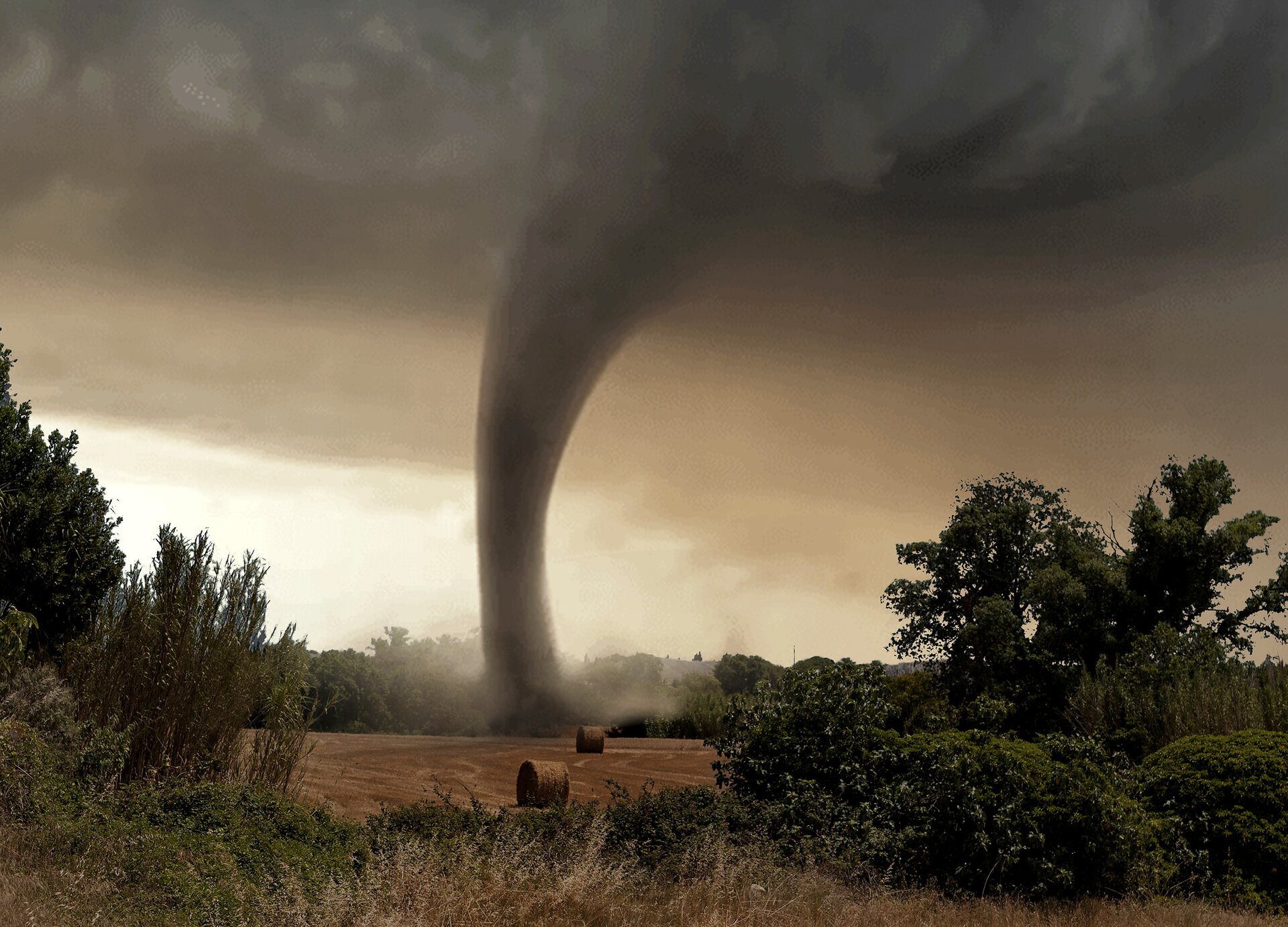 Zdjęcie przedstawia trąbę powietrzną znajdująca się nad polem otoczonym drzewami. Trąba ma kolor ciemnoszary. W miejscu, w którym znajduje się trąba powietrza, widoczna jest uskakująca na boki ziemia. Niebo jest siwo‑czarne, z głębiącymi się stalowymi chmurami. Na polu widoczne są bale ze słomą.