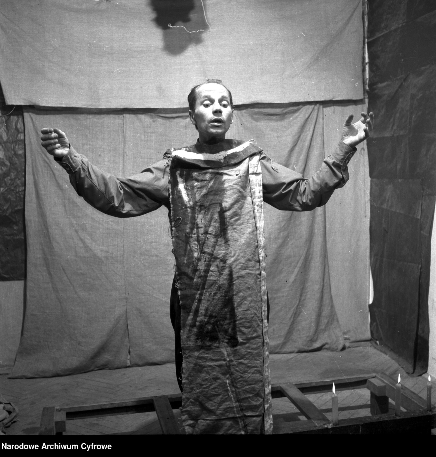 Obraz przedstawia dojrzałego mężczyznę stojącego ma tle płachty materiału. Mężczyzna ma rozłożone szeroko ręce. Ubrany jest w koszulę i spodnie. Na ramiona ma zarzuconą pogniecioną tkaninę, która sięga mu aż do kostek. Sprawia to wrażenie, jakby mężczyzna ubrany był w worek.