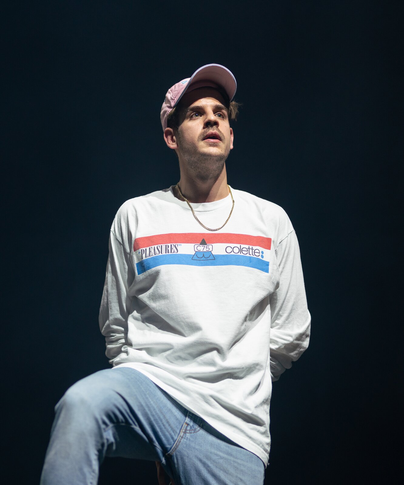 Zdjęcie przedstawia Taco Hemingway'a [czytaj: tako hemingłeja]. Młody mężczyzna stoi na scenie, oświetlony reflektorem. Ręce trzyma za sobą, a kolano prawej nogi ma uniesione do góry. Ma na sobie dżinsy i białą koszulkę z długim rękawem. Na głowie ma czapkę z daszkiem. Ma niewielki zarost, patrzy przed siebie. Tło jest czarne.