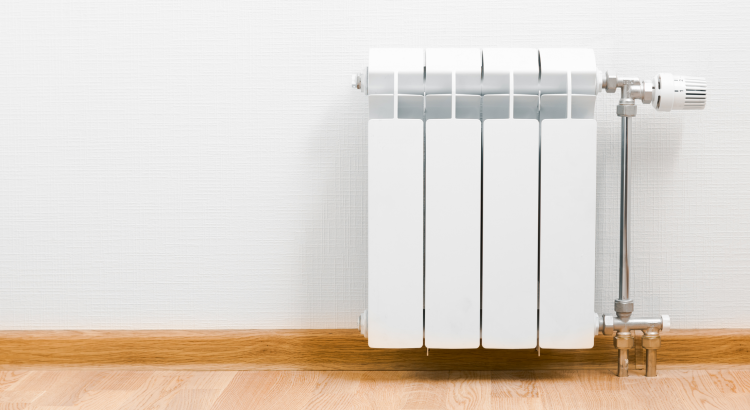 Na zdjęciu widoczny jest biały grzejnik ścienny zawieszony na ścianie. Na prawo widoczne są przewody podłączone do grzejnika oraz pokrętło termostatu.