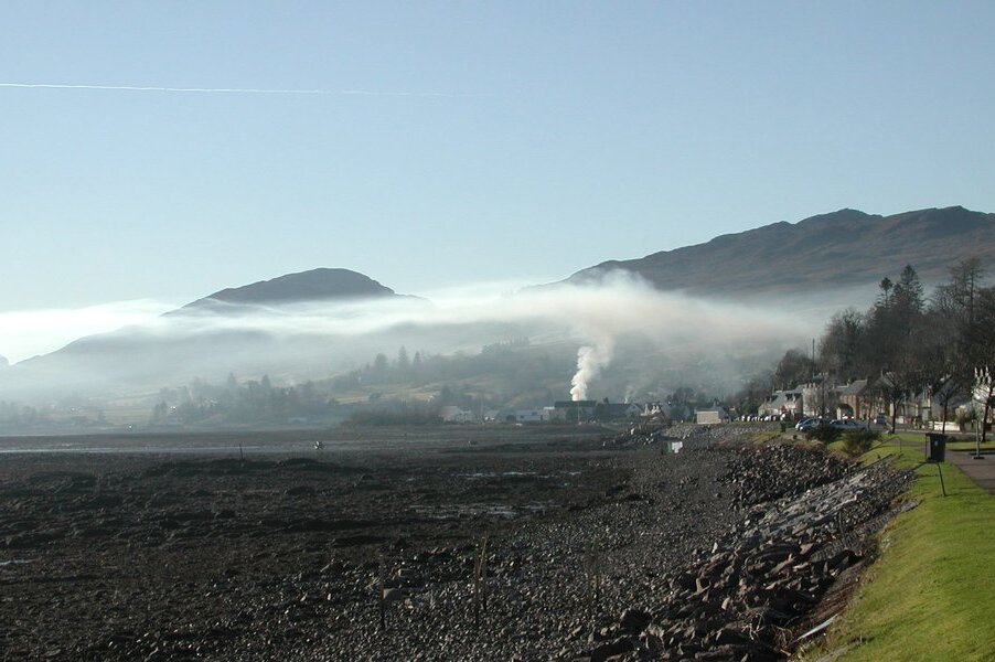 Fotografia barwna. Ilustracja przedstawia krajobraz górski. Zdjęcie prezentuje również pole uprawne oraz wieś. Z komina jednego z domów unosi się dym. Dym rozchodzi się po całej dolinie.