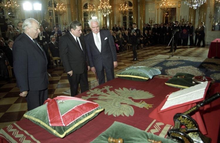 Zdjęcie przedstawia uroczystość państwową na Zamku Królewskim w Warszawie. W dużej sali, na stole z rozłożonym starym Proporcem Prezydenta Rzeczpospolitej Polskiej leżą na aksamitnych poduszkach Order Orła Białego i Order Odrodzenia Polski. Przed insygniami stoi Prezydent Lech Wałęsa oraz ostatni Prezydent na Uchodźctwie, Ryszard Kaczorowski. W sali są także licznie zgromadzeni politycy i obserwatorzy.