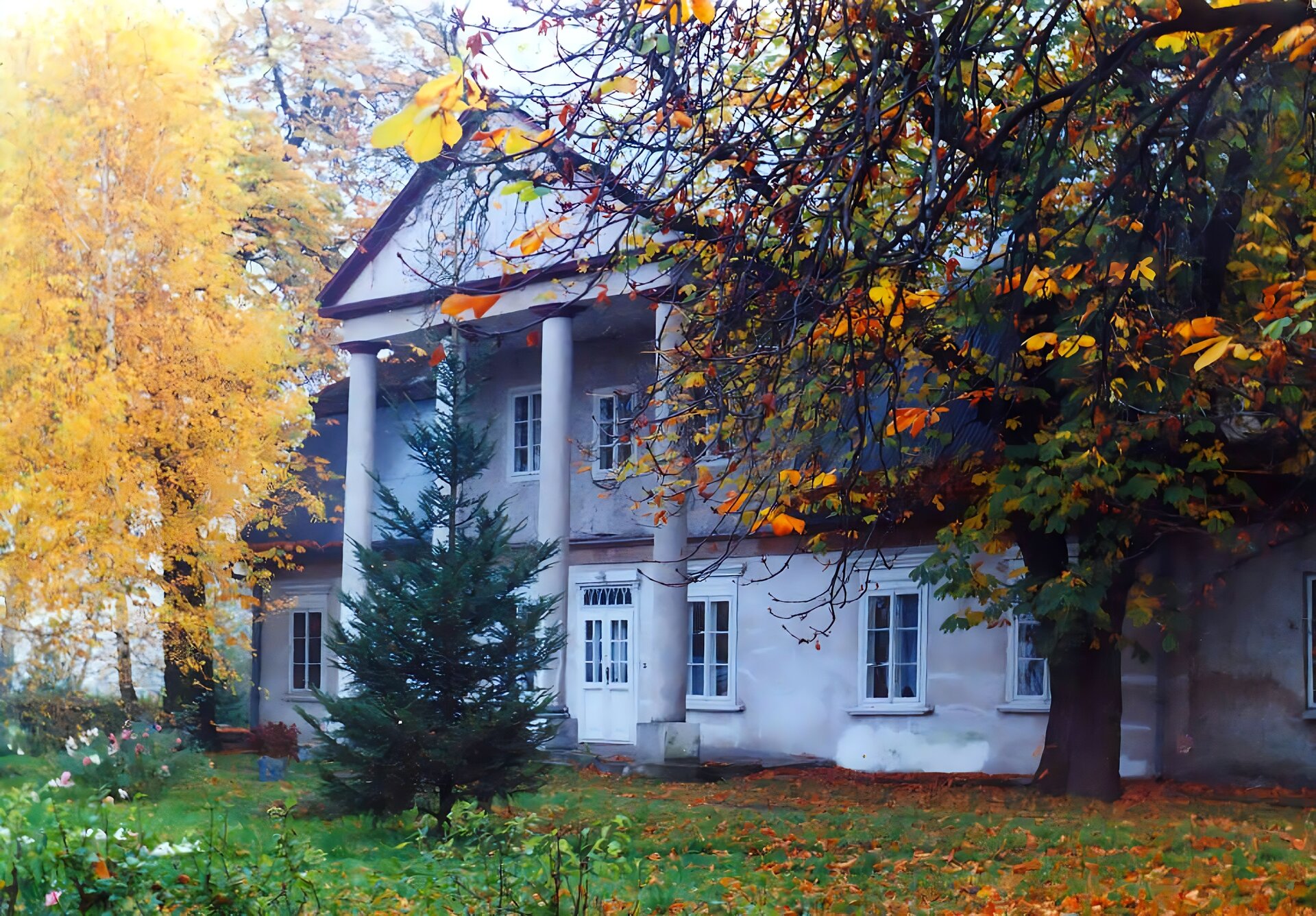Zdjęcie przedstawia biały, piętrowy dworek. Nad wejściem znajduje się wysunięty dach, który podparty został czterema kolumnami. Okna rozmieszczone są symetryczne. Wokół rosną wysokie drzewa z żółtymi i czerwonymi liśćmi Przed dworkiem rośnie pojedyncza choinka, a wokół zielona, niska trawa. Zdjęcie zostało wykonane w świetle dziennym.