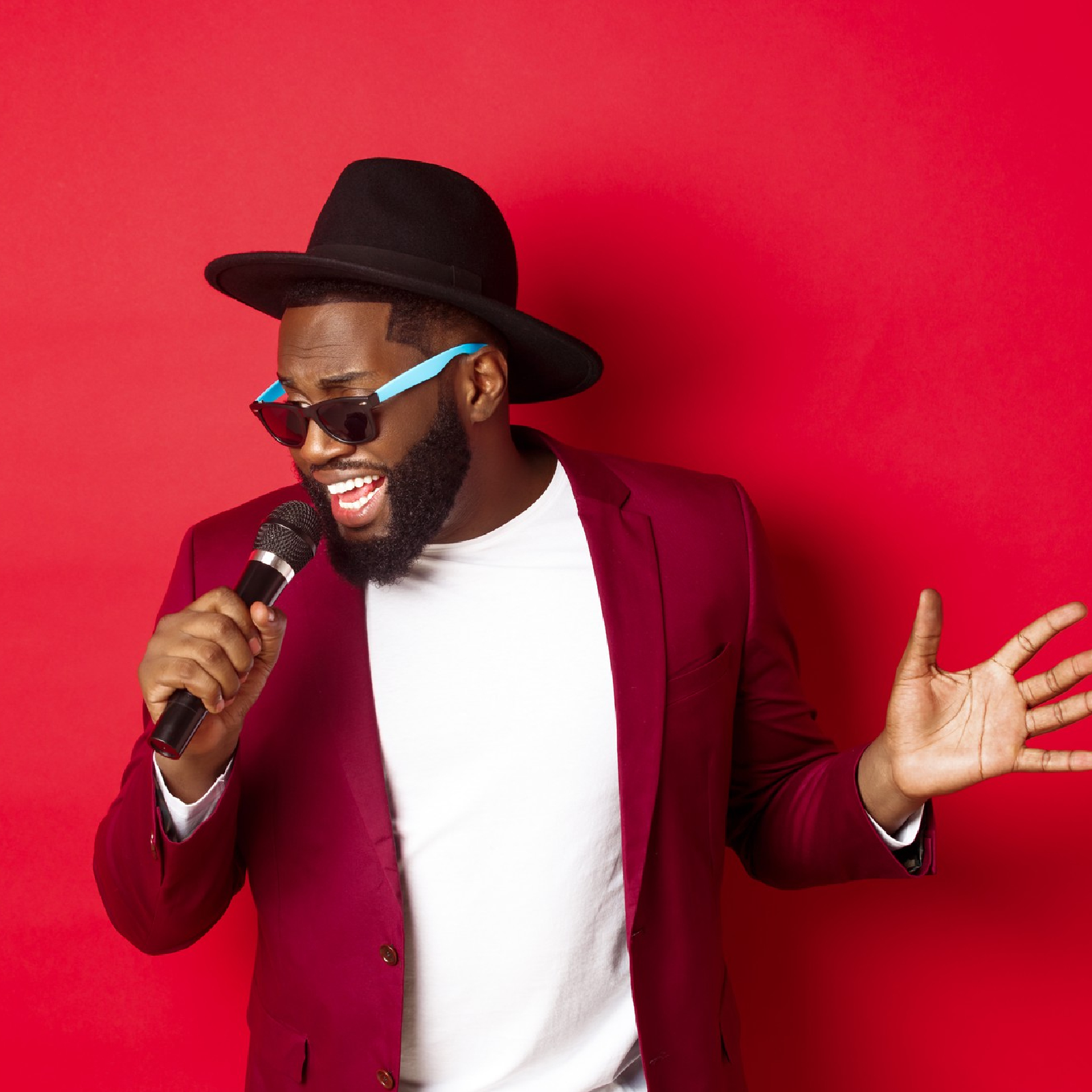 Ilustracja przedstawia czarnoskórego mężczyznę w kapeluszu i okularach przeciwsłonecznych, śpiewającego do mikrofonu. Mężczyzna ma zadbany zarost, nosi czerwoną marynarkę z białym podkoszulkiem. 