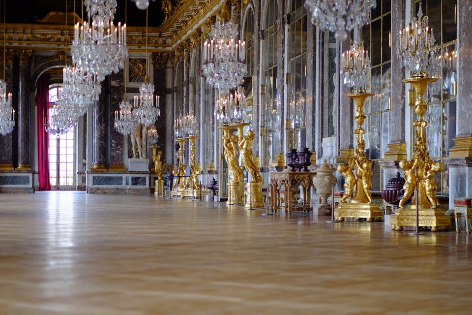 Na zdjęciu znajduje się fragment wnętrza pałacowej sali. Po prawej stronie są umieszczone wysokie lustra w złotych ramach, zakończone łukami. Przed lustrami, w rzędzie stoją ogromne złote lichtarze, w których znajdują się kryształowe ozdoby i świece. Lichtarze trzymają figury kobiet i dzieci. Pomiędzy lichtarzami stoją wazy. Na wprost znajdują się oszklone, wysokie drzwi, prowadzące na balkon, zwieńczone łukiem, w którym wisi bordowa zasłona. Po obu stronach drzwi są kolumny. W rogu stoi rzeźba nagiego mężczyzny. Na suficie wiszą kryształowe żyrandole, na których znajdują się dwa rzędy świec. Na ścianach, przy suficie znajdują się złoto rzeźbione gzymsy oraz pilastry. Podłoga jest drewniana.