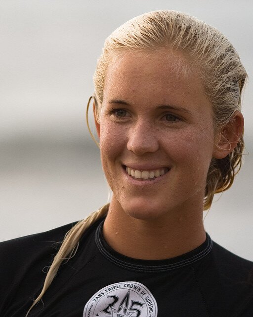 Fotografia młodej dziewczyny w piance surferskiej. Ma jasne włosy, uśmiecha się.