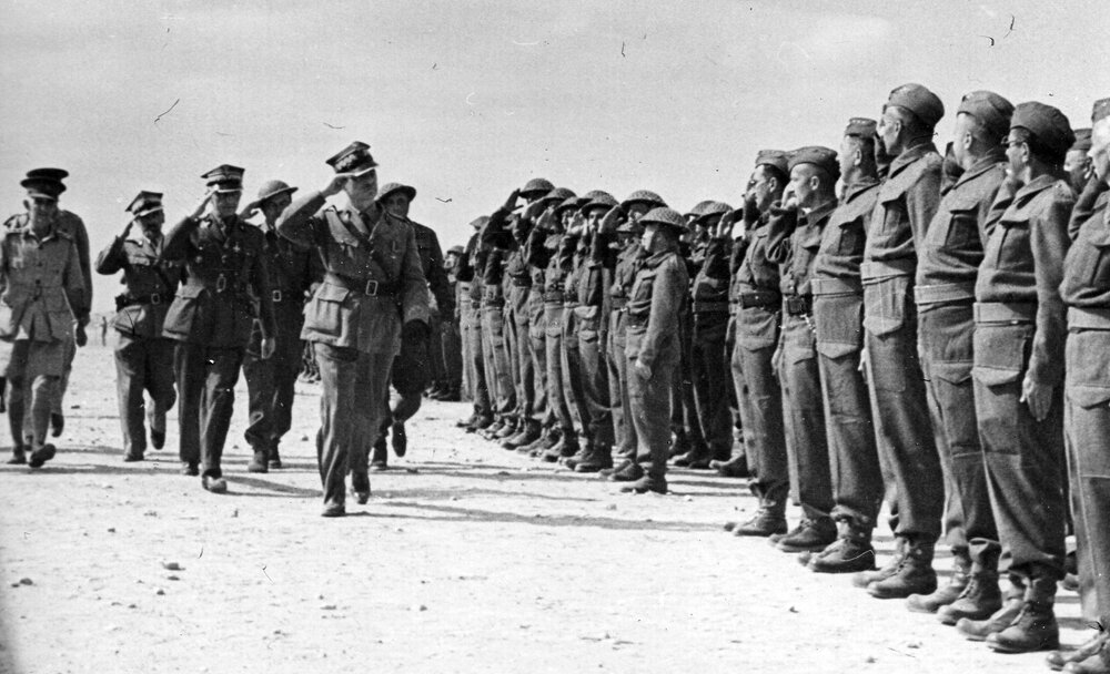 Zdjęcie przedstawia żołnierzy. Stoją na baczność w szeregach. Wzdłuż nich idzie generał. Salutuje lewą dłonią do czapki. Za nimi idą oficerowie. 
