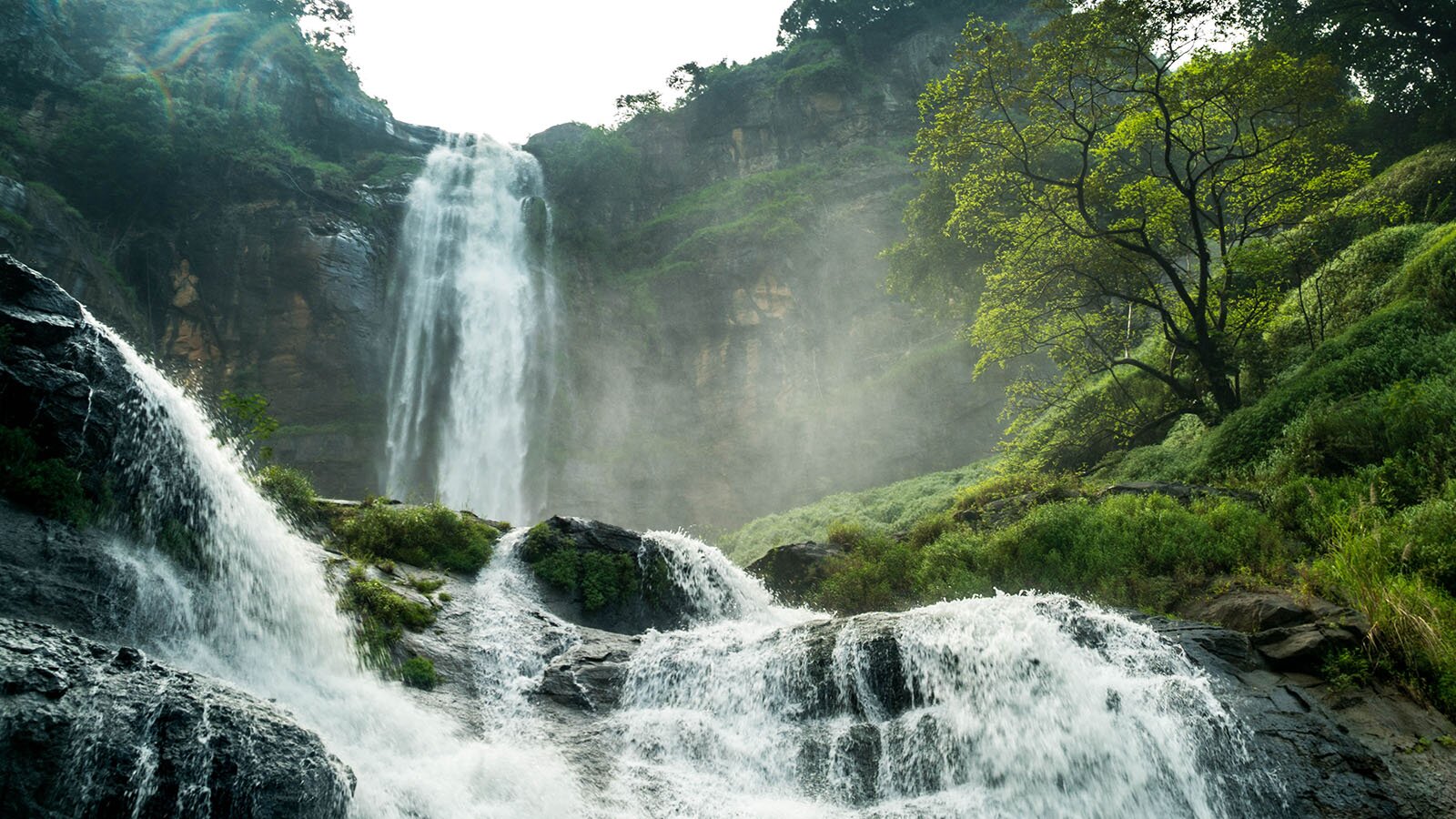 Zdjęcie przedstawia dwukaskadowy wodospad. Ujęcie wykonane z dołu ukazuje pierwszą cześć wodospadu lejącego się z wysokiego klifu oraz niżej mniej stromą część, gdzie woda spływa kaskadowo po skałach.