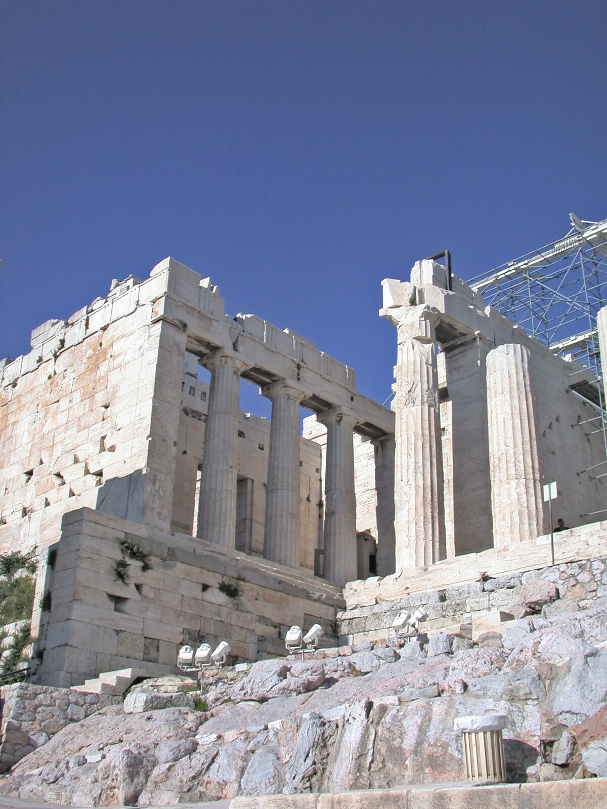 Fotografia przedstawia pozostałości po starożytnym budynku, który został wykonany z jasnego kamienia. Budynek konstrukcyjnie został oparty na kolumnach, które podtrzymują część ścian. Część budynku poddana została renowacji o czym świadczyć może rusztowanie.