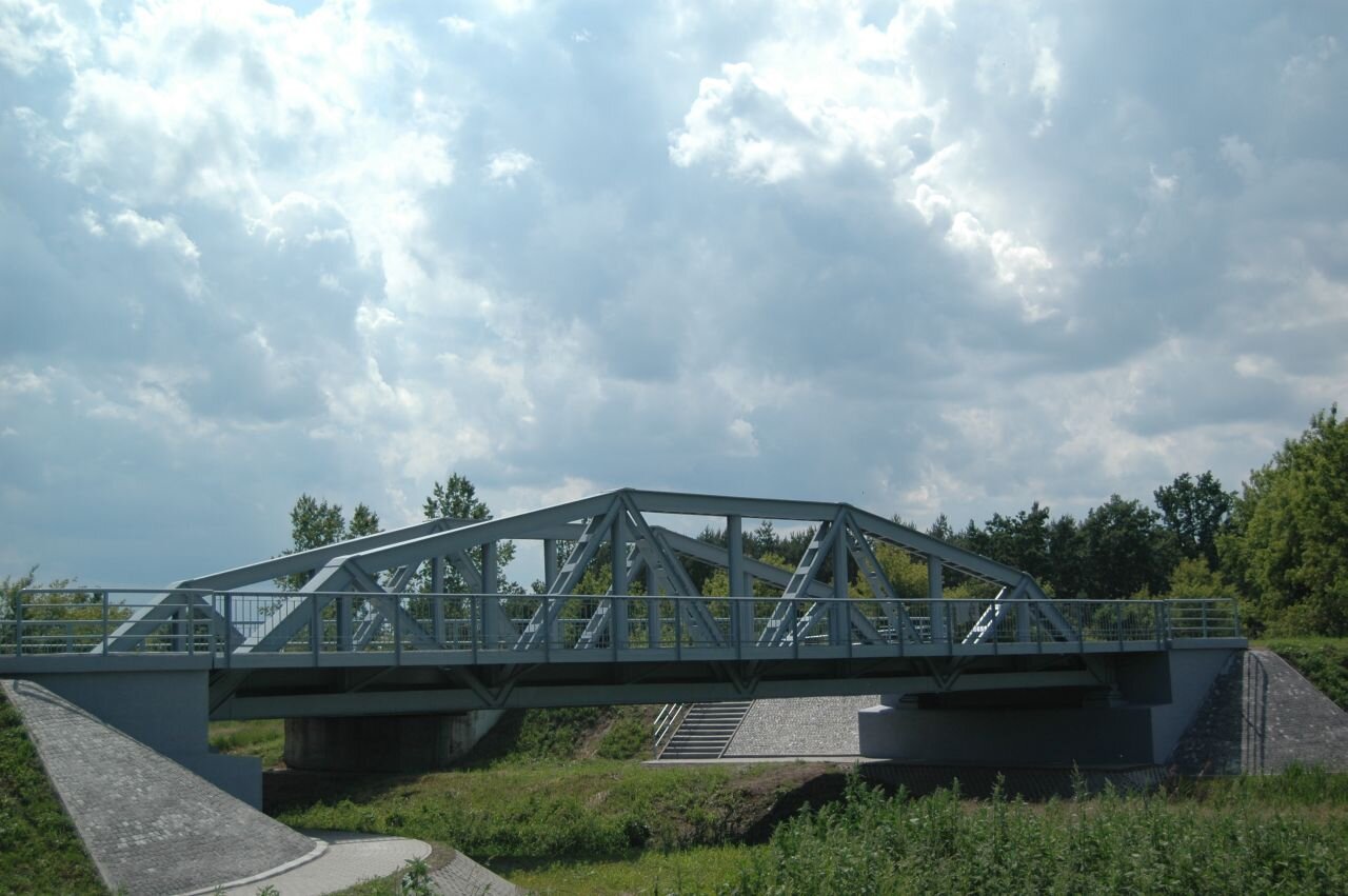 Ilustracja przedstawia most o konstrukcji łukowej, gdzie powierzchnia jezdna jest umiejscowiona u dołu. Konstrukcja stalowa oparta jest na szerokich podporach skrajnych, zwanych przyczółkami. Stalowa konstrukcja mostu została pomalowana na biały kolor, a przyczółki obłożone są betonową kostką.