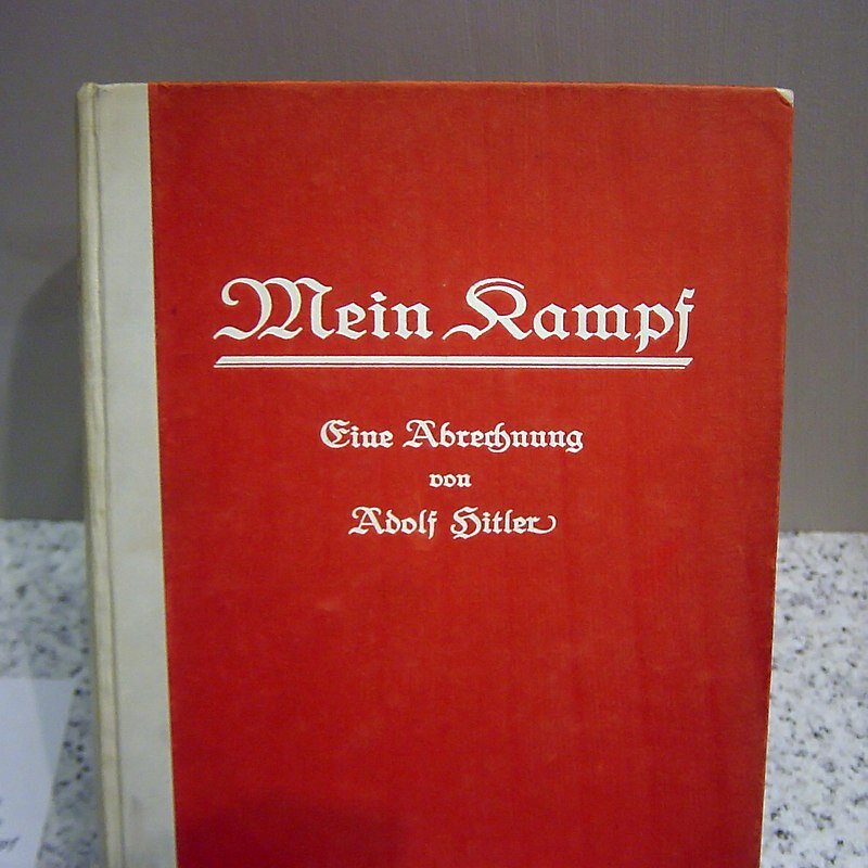 Zdjęcie przedstawia oprawioną w czerwona obwolutę książkę. Tytuł zapisany ozdobną, złotą czcionką: Mein Kampf. poniżej Eine Abrechnung von Adolf Hitler. 