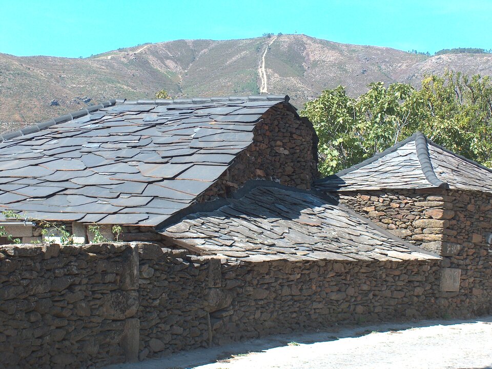 Na zdjęciu jest kamienny mur i kamienna zabudowa, włącznie z dachem. W tle są wzniesienia. Na fotografii jest bezchmurne niebo. 