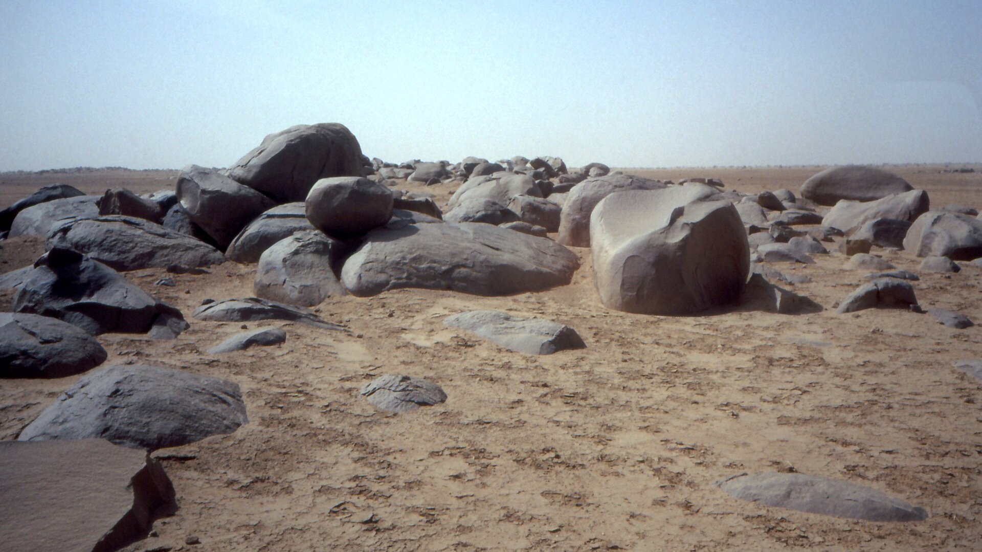 Zdjęcie przedstawia pustynię na Płaskowyżu Al‑Hamada al‑Hamra, centralna Sahara. Na zdjęciu duże, okrągłe kamienie na piasku. Mają ciemnoszary kolor i najczęściej obłe kształty. Równy, rozległy teren. Niebieskie, bezchmurne niebo.