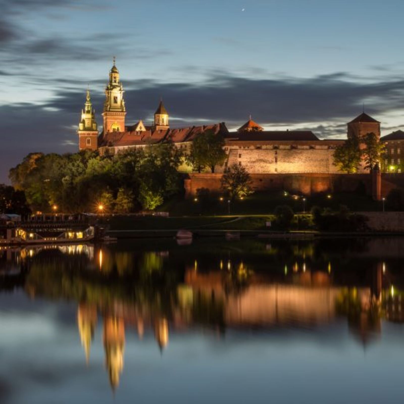 Zdjęcie przedstawia wieczorne ujęcie Zamku i Katedry na Wawelu, odbijające się w wodach Wisły. Oświetlony zabytek dostojnie wygląda na wzgórzu wawelskim.