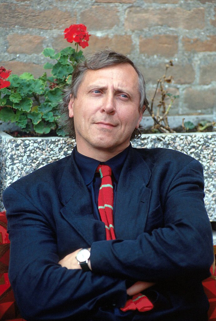 Zdjęcie przedstawia dojrzałego mężczyznę. Ma on owalną twarz, długi nos, małe oczy. Włosy szpakowate, proste. Ubrany jest w garnitur, koszulę i krawat. Mężczyzna siedzi z założonymi rękami. Za jego plecami gazon z kwitnącymi pelargoniami.