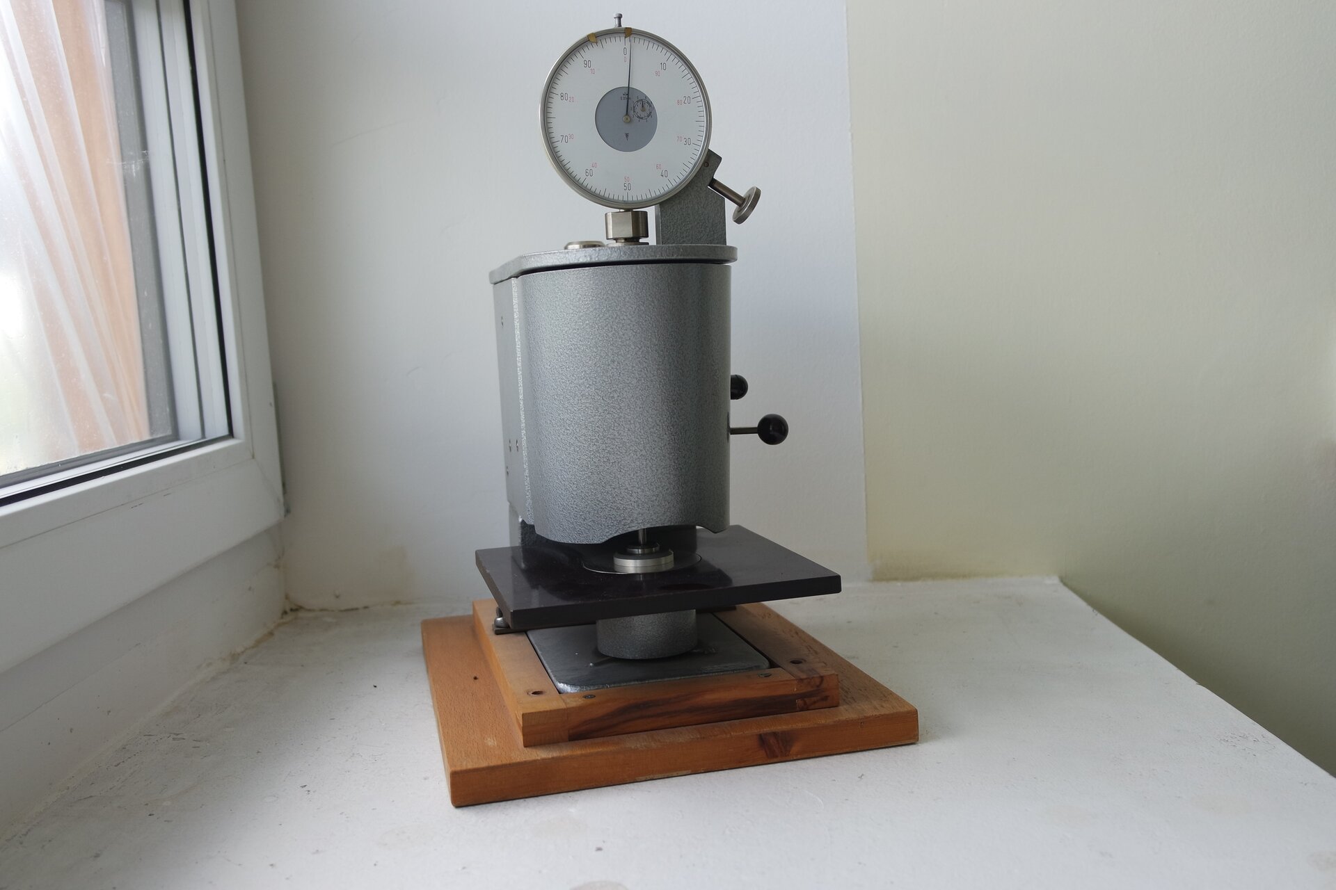 Zdjęcie prezentuje grubościomierz mechaniczny do pomiaru grubości. Urządzenie, stojące na drewnianej stopce, zbudowane jest z układu pomiarowego, układu zerującego i czujnika zegarowego pokazującego wynik pomiaru.