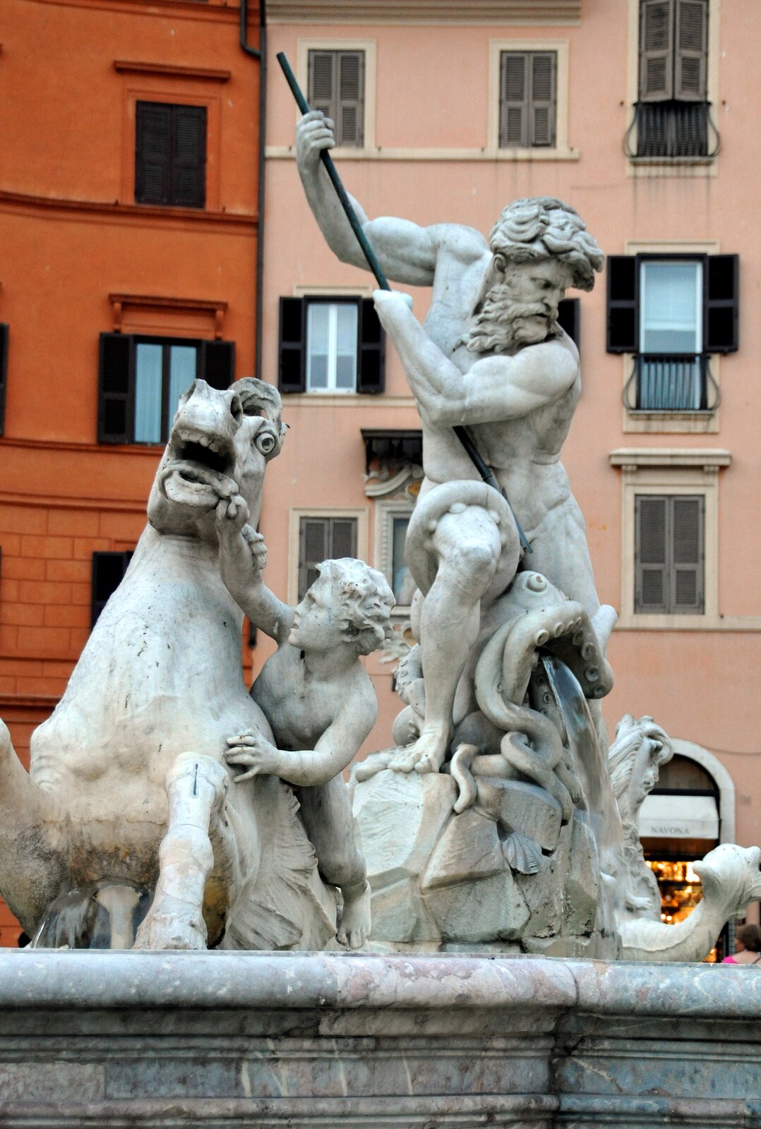 Ilustracja przedstawia fragment fontanny znajdującej się na Piazza Navona w Rzymie. Figury wykonane są z białego marmuru. W centrum fotografii widnieje postać Neptuna w otoczeniu morskich stworów. Bóg mórz wbija trójząb w łeb ośmiornicy, która otacza go mackami. Z paszczy ośmiornicy leje się woda. Na lewo znajduje się figura przypominająca konia, którego stara się okiełznać mały chłopiec.