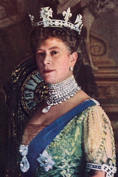 Zdjęcie przedstawia kobietę. Ma krótkie, ciemne, kręcone włosy oraz srebrną koronę na głowie. Ma duży, wysadzany diamentami naszyjnik. Ubrana jest w suknię przepasaną niebieską szarfą. 