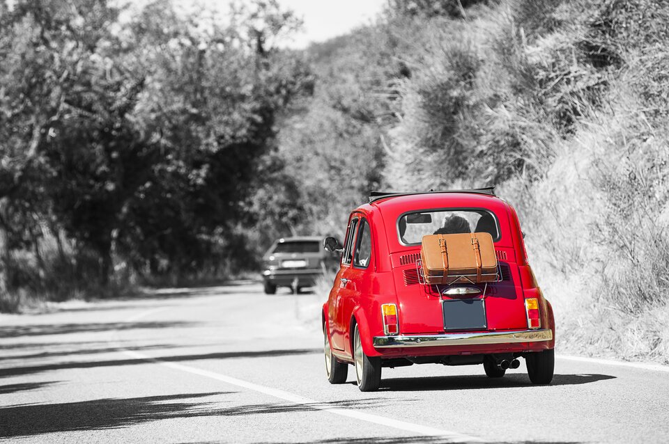 Zdjęcie przedstawia klasyczny, czerwony Fiat 500 z brązową skórzaną walizką przypiętą do tylnej części. Samochód jedzie krętą, wiejską drogą. Auto wyróżnia się żywym kolorem, podczas gdy reszta scenerii – drzewa, trawa i inny samochód w oddali – jest w czarno-białych odcieniach. Kierowca jest widoczny przez tylne okno.