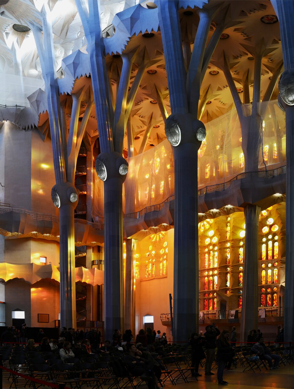 Ilustracja przedstawia wnętrze katedry Sagrada Família. Ukazuje wysokie kolumny przypominające pnie drzew, które rozgałęziają się ku górze i podtrzymują skomplikowane, geometryczne sklepienie. Światło wpadające przez kolorowe witraże tworzy na ścianach i posadzce ciepłe odcienie żółci i złota. W dolnej części widoczne są rzędy krzeseł oraz zwiedzający.